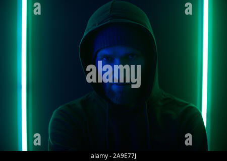 Portrait of young bearded man in dark clothing wearing hoody standing against the wall with green light Stock Photo