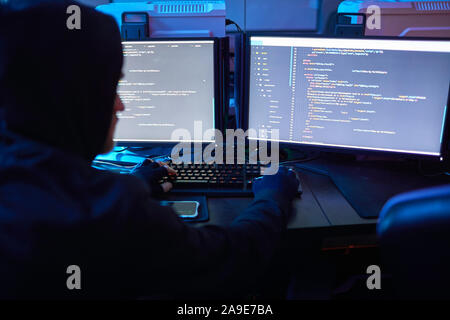 Rear view of computer hacker wearing hoody sitting at the table in front of computer monitors and breaking the computer codes in dark office Stock Photo