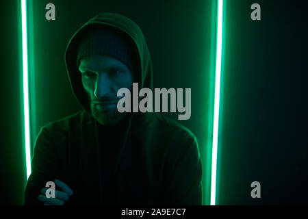 Young bearded man wearing black hoody shirt standing with arms crossed against the wall with green light Stock Photo