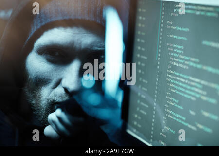 Portrait of young bearded computer programmer looking at camera near the computer monitor with software in dark office Stock Photo