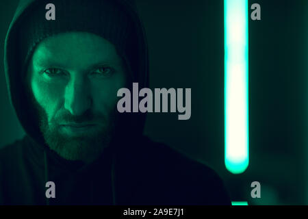 Portrait of serious bearded gangster wearing black hoody shirt looking at camera with green lights in the background Stock Photo