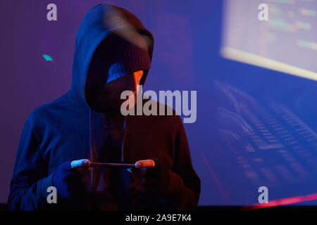 Bearded computer hacker in black hoody shirt holding smart phone and working online in dark room Stock Photo