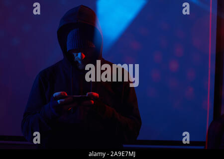 Image of computer hacker in black clothing standing and using his mobile phone in dark room Stock Photo