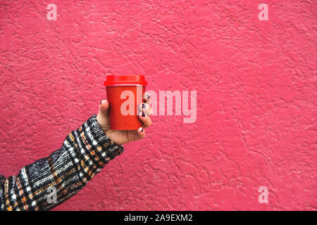 A red cup of take away coffee in a female hand in an autumn coat against a bright burgundy wall. Background and copy space. Stock Photo