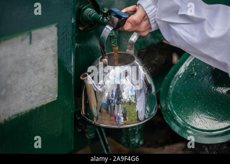 A metal kettle in hands under a stream of water. Hot tea pours from the tap. Volunteer makes a drink. Field army kitchen on the street. Hot water in t Stock Photo