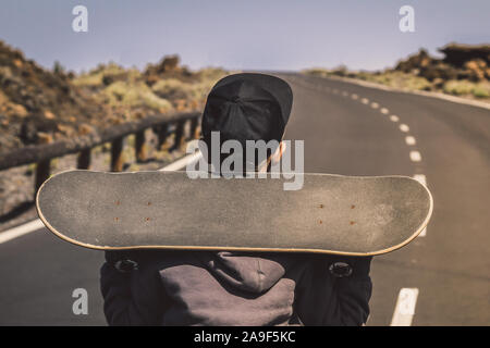Hipster trendy boy holding skateboard on shoulder walking in the middle of the mountain road. Close up back view of a beautiful kid with skate enjoyin Stock Photo