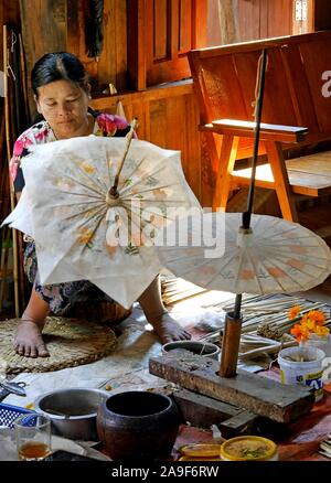 Pretty young Burmese woman skilfully painting handmade paper parasol surrounded by paints and brushes. In stilt house on Inle Lake, Myanmar Stock Photo
