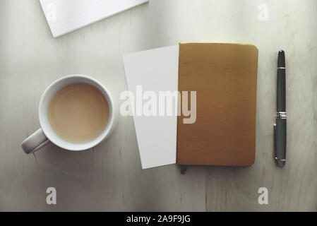 Top view of a mug with coffee, a notebook, a pen and blank paper on a wooden desk table next to a window. Empty copy space for Editor's text. Stock Photo