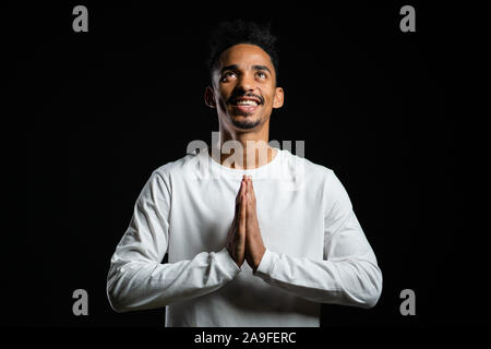 Handsome african american man in white t-shirt praying over black background. Mixed race guy begging someone. Stock Photo