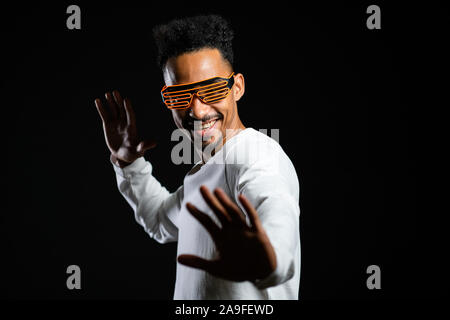 African man in glowing neon glasses moving to the music rhythm on black studio background. Dancing positive comic guy.  Stock Photo