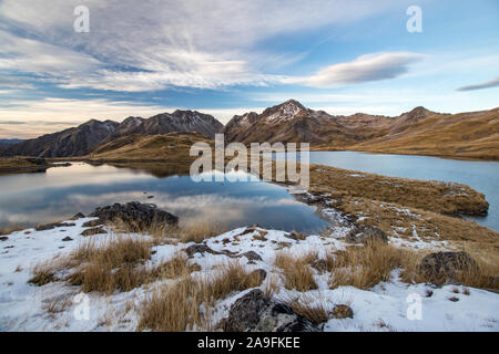 Angelus lakes mirroring fine clouds and blue sky after sunset. Rocky mountains in background with snow, tussock. Angelus mountain hut in the middle