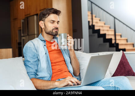 Young adult man sitting at home, using laptop, drinking beverage Stock Photo