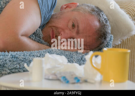 Man suffering from flu or cold lies sadly covered with a blanket on his bed. Drugs, medicines, cup and tissues in foreground Stock Photo