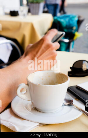closeup of a young caucasian man having a cappuccino, while using his smartphone, sitting at a table in an outdoors terrace of a coffeehouse Stock Photo