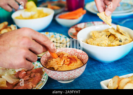 some people having some different appetizers, such as hummus, potato chips and salmon and cod carpaccio, served on different colorful plates and bowls Stock Photo