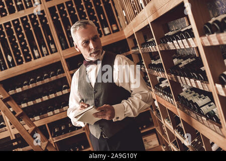 Sommelier Concept. Senior man standing taking notes on wine collection thoughtful Stock Photo