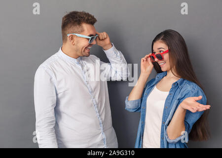 Relationship Concept. Young couple studio standing isolated on grey taking off glasses looking at each other happy Stock Photo