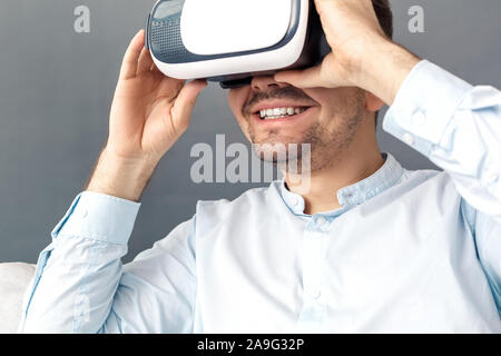 Freestyle. Young man looking through virtual reality headset studio sitting on sofa isolated on grey smiling excited close-up Stock Photo