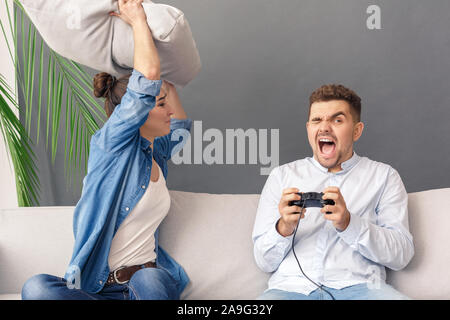 Relationnship Concept. Young couple woman hitting with pillow man playing game console screaming sitting on sofa studio isolated on grey Stock Photo