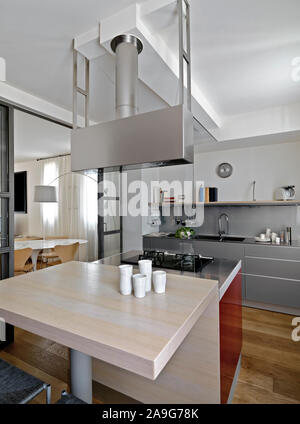 interior shot of a modern kitchen in foreground the kitchen island with wood dining table and gas hob while on background the kitchen cabinets with bu