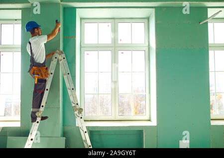 A builder standing on a ladder installs drywall at a construction site Stock Photo