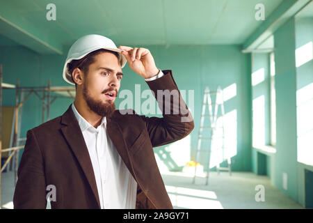 Builder foreman in a white helmet surprised in shock at a construction site indoors Stock Photo
