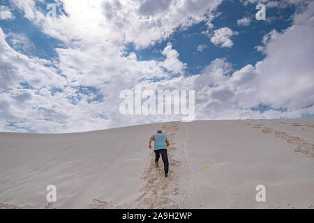 man in casual clothing running up a white sand dune against blue sky Stock Photo