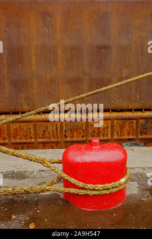 Rope from a ship tied to an old red concrete mooring bollard on a quayside. Stock Photo