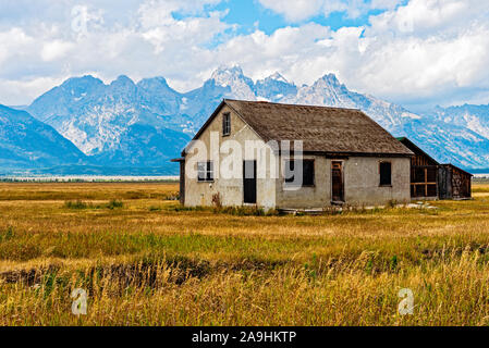 Abandoned pioneer farm home in grassy field with tall Rocky Mountains beyond under blue skies with white fluffy clouds. Stock Photo
