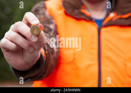 a person wearing cammo and hunter orange holds a 12 gauge shotgun shell in front of them in the forest Stock Photo