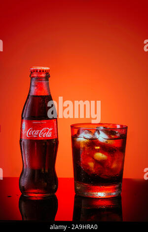 Italy, Milan - 15 November 2019: Classic bottle Of Coca-Cola and glass of fresh Coca Cola with ice cubes and water drops on mirror surface. Studio shot. Red background Stock Photo