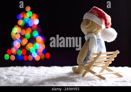 Brown teddy bear wearing scarf and santa hat sitting on wooden bench with snow in winter looking at colorful bokeh lights of Christmas tree. Stock Photo