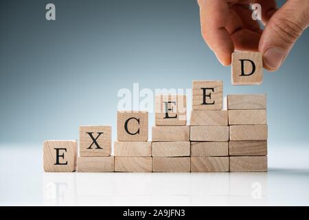 Person's Hand Placing Exceed Text Over Stack Of Wooden Blocks In A Row Stock Photo