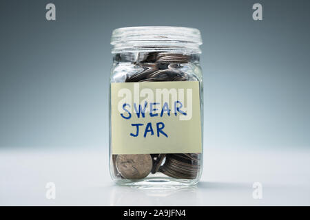 Close-up Of A Glass Jar With Swear Jar Text Filled With Coins Against Grey Background Stock Photo