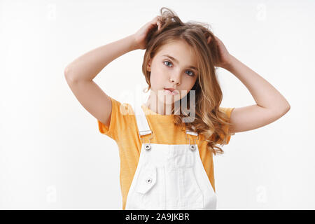 Tender lovely young blond girl in summer dungarees over yellow t-shirt, look thoughtful and cute camera, scratch hair bothered, touch hairstyle, suffe Stock Photo