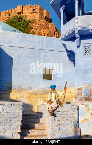 Elderly man in a blue turban sitting on a wall in the Blue City of Jodhpur with Mehrangarh Fort in the background, Rajasthan, India Stock Photo