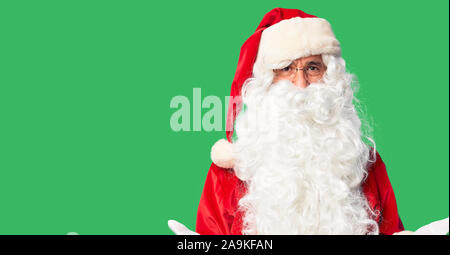 Middle age handsome man wearing Santa Claus costume and beard standing smiling cheerful with open arms as friendly welcome, positive and confident gre Stock Photo