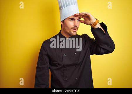 Young chef man wearing uniform and hat standing over isolated yellow background very happy and smiling looking far away with hand over head. Searching Stock Photo