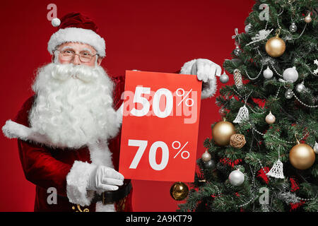Portrait of senior man in Santa costume holding placard with big sale while standing near the Christmas tree over red background Stock Photo