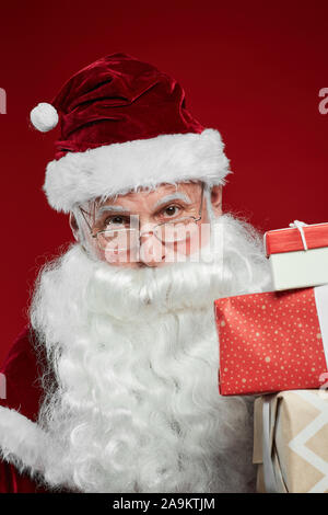 Portrait of Santa Claus in hat holding gift boxes and looking at camera isolated on red background Stock Photo