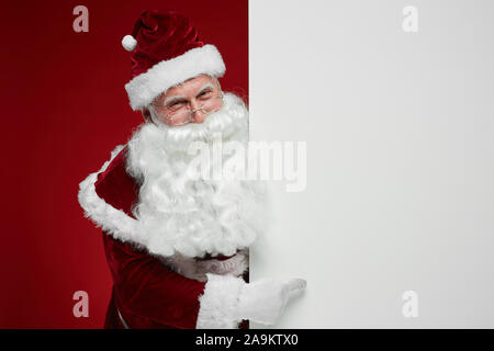 Portrait of Santa Claus standing near the big billboard and pointing at it isolated on red background Stock Photo