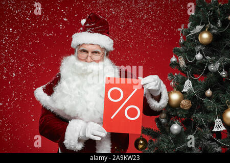 Portrait of Santa Claus holding sale sign and showing it to the camera while standing near the Christmas tree during snowing Stock Photo