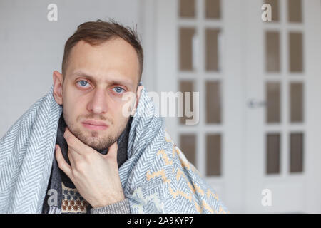 Young man with scarf, covered his head with a blanket, holds his neck because of sore throat Stock Photo