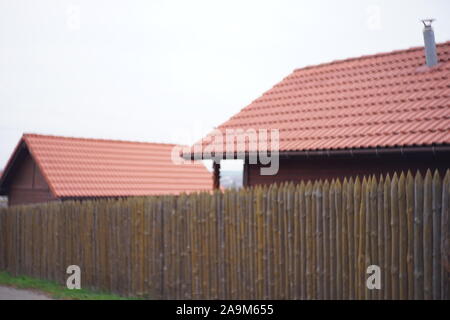 Fence made of sharp wooden stakes. Wooden houses with red tiled roof and sky. Unfocused image. Stock Photo