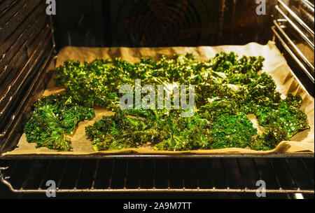 Freshly made crispy kale chips on a baking tray and baking paper in a lit up oven with open oven door - angled view, horizontal landscape format Stock Photo