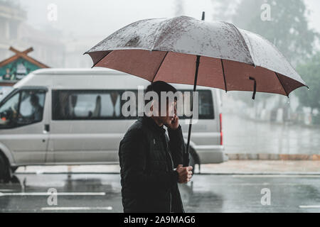 Sapa, Vietnam - 13th October 2019: A middle aged asian man walks past the camera in the rain under an umbrella in the small mountain town of Sapa Stock Photo