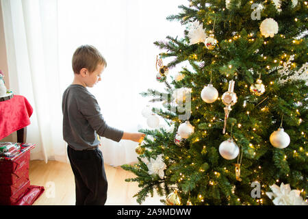 Young boy stands placing ornament on Christmas tree during holiday Stock Photo