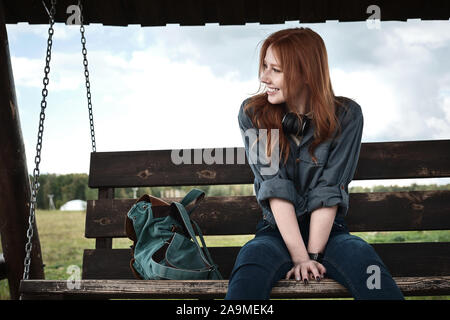Redhead girl in a jeans shirt sits with a backpack on a wooden swing bench, leaning forward and smiling, looking to the side. Stock Photo
