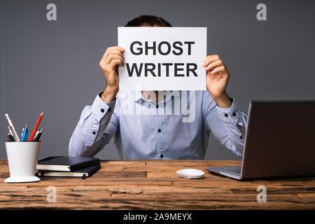 Businessman Hiding His Face Behind Card With Ghost Writer Text Sitting In Front Of Desk Stock Photo