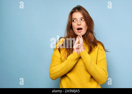 The brunette stands on a blue background in a yellow sweater and looks away, opening her eyes and mouth wide Stock Photo